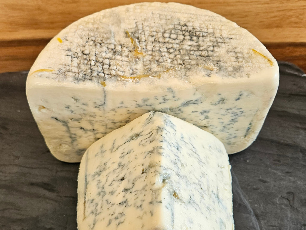 A close-up of a wedge of blue cheese with a white rind and blue-green marbling, with a smaller cut piece in front, set on a dark slate surface with a wooden background.