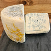 Two wedges of blue cheese on a dark slate board with a wooden background. One wedge shows the white rind with orange zest, and the other shows the creamy off-white interior with blue mold veins.